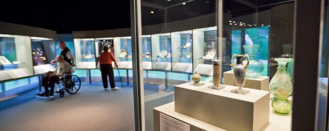 Ancient glass bottles are in a glass case. A man stands behind a person in a wheelchair and a woman with a cane are out of focus in the background.