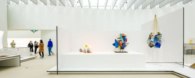 Wide shot of a white walled gallery with colorful glass sculptures and people in the distance in the far left
