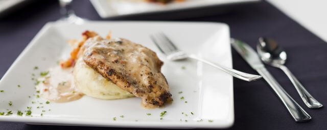A roasted chicken breast sits atop a bed of creamy mashed potatoes on a plate garnished with chopped parsley and a fork nearby
